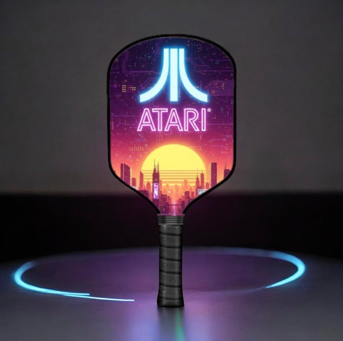 A retro-futuristic Atari-branded paddle-shaped light-up sign or lamp stands upright on a dark surface. The front has a neon-style Atari logo and the word ATARI over a colorful sunset cityscape, with a glowing blue circular ring beneath it.