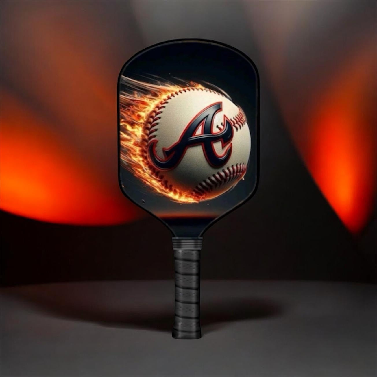 A pickleball paddle featuring a printed graphic of a baseball engulfed in flames, with the Atlanta Braves 'A' logo on the paddle face, set against a dark background with dramatic orange lighting.
