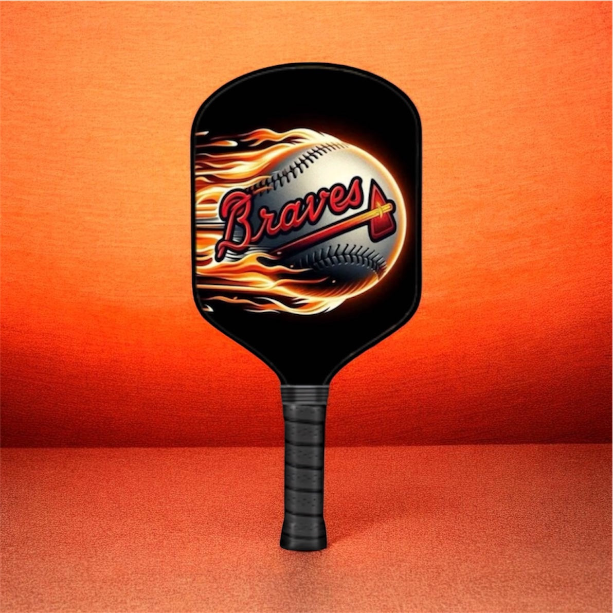 A black pickleball paddle featuring a graphic of a baseball engulfed in flames with the word 'Braves' in red script, set against a textured orange background.