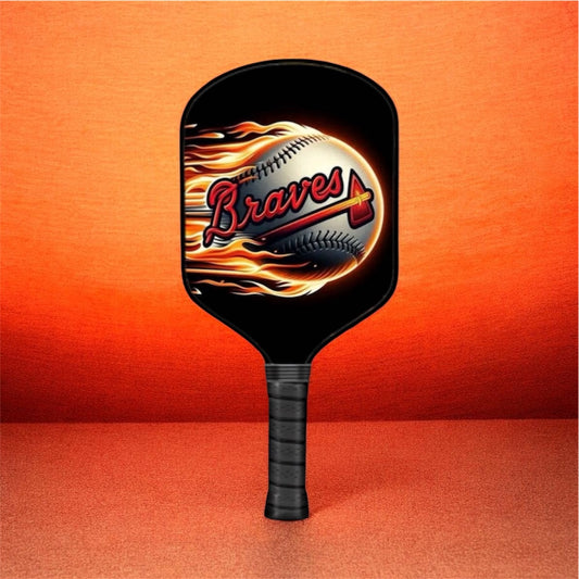 A black pickleball paddle featuring a graphic of a baseball engulfed in flames with the word 'Braves' in red script, set against a textured orange background.