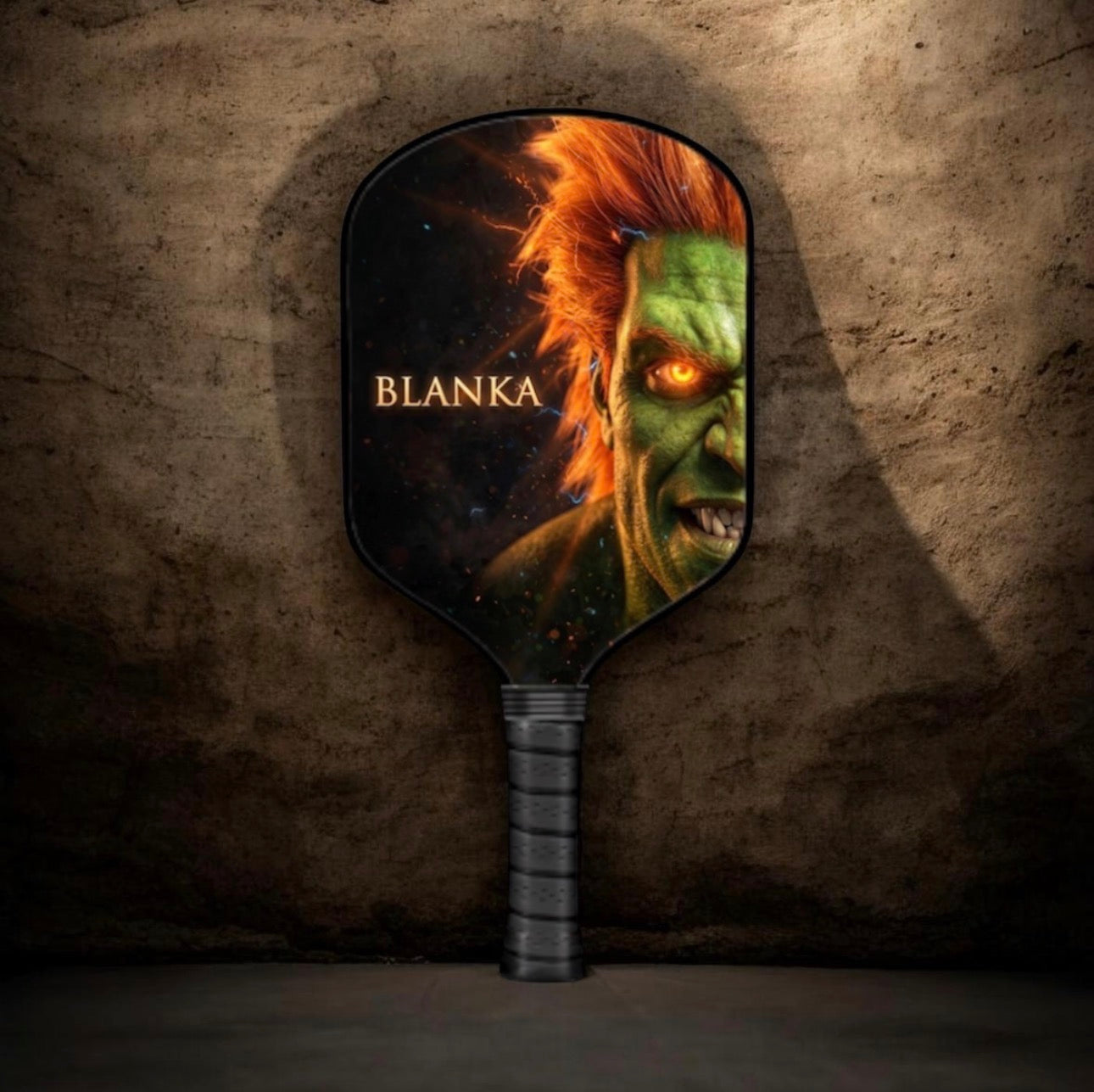 A paddle-shaped mirror with a dark handle, featuring a printed fantasy creature face with the name 'BLANKA' on the face, set against a textured dark brown stone wall background.