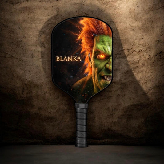 A paddle-shaped mirror with a dark handle, featuring a printed fantasy creature face with the name 'BLANKA' on the face, set against a textured dark brown stone wall background.