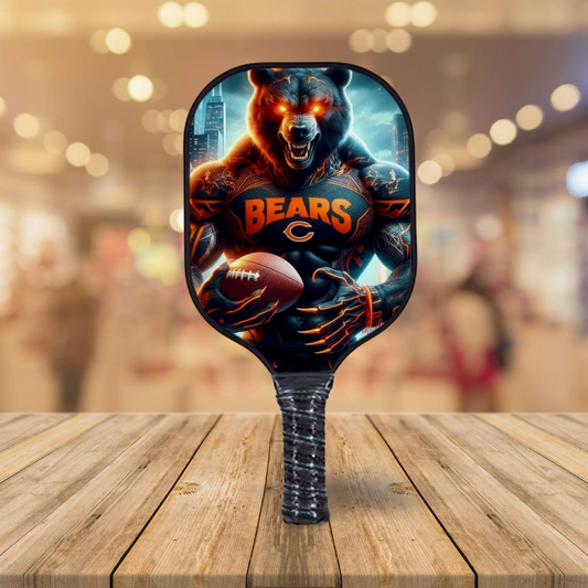 A pickleball paddle with a Chicago Bears themed design featuring a bear holding a football, positioned on a wooden surface with a blurred shopping mall background.
