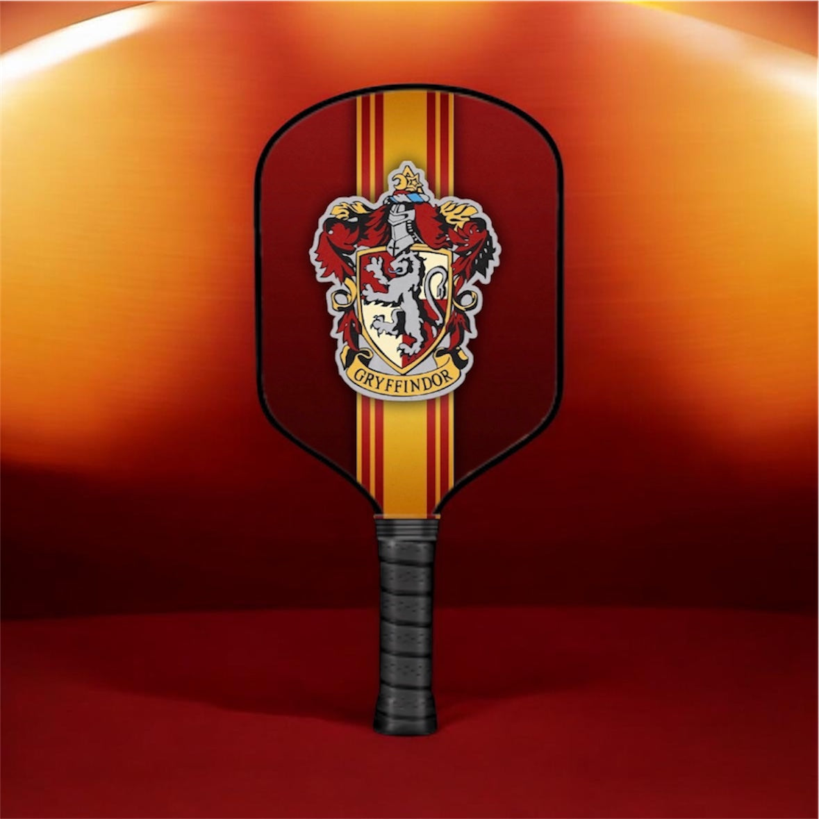 A Gryffindor-themed pickleball paddle with a red and gold striped face, featuring the Gryffindor crest in the center, set against a warm orange and red gradient background.