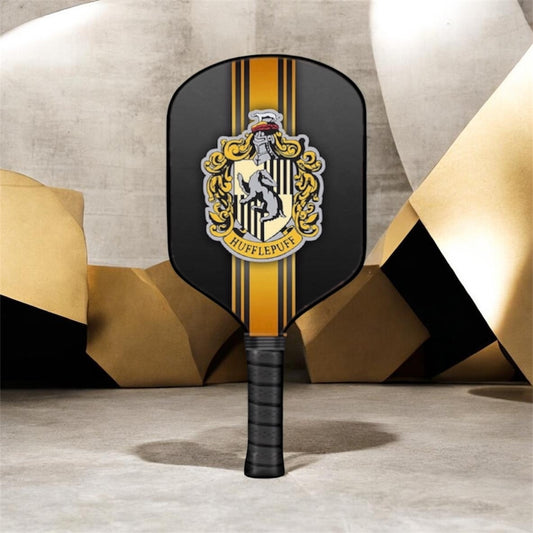 A pickleball paddle with a black face featuring a gold and yellow vertical stripe design and a Hufflepuff crest, standing upright against a modern geometric background.
