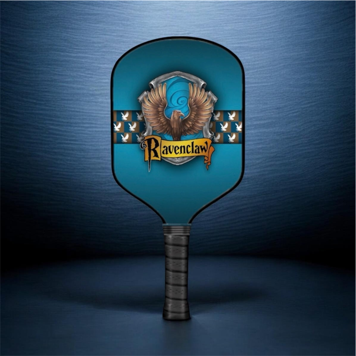 A Ravenclaw-themed paddle-shaped item, likely a fan or decorative prop, featuring the Slytherin crest with an eagle and the word 'Ravenclaw' on a blue background with a dark textured studio backdrop.