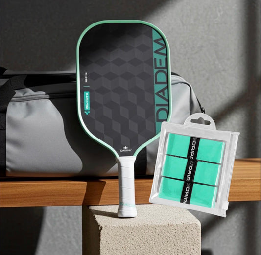 A Diadem pickleball paddle stands upright on a stone block beside a gray bag, with a packaged set of teal grip tape in front. The paddle has a mint green border and dark gray geometric face, and the grip tape pack is labeled Diadem Grip Tape.