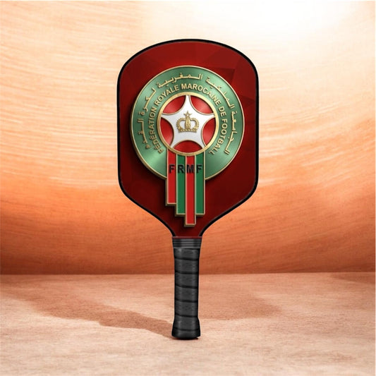 A Moroccan national futsal paddle with a dark red head, featuring the Moroccan Football Federation emblem in green, gold, and red, and a black wrapped handle, set against a warm wood-grain background.