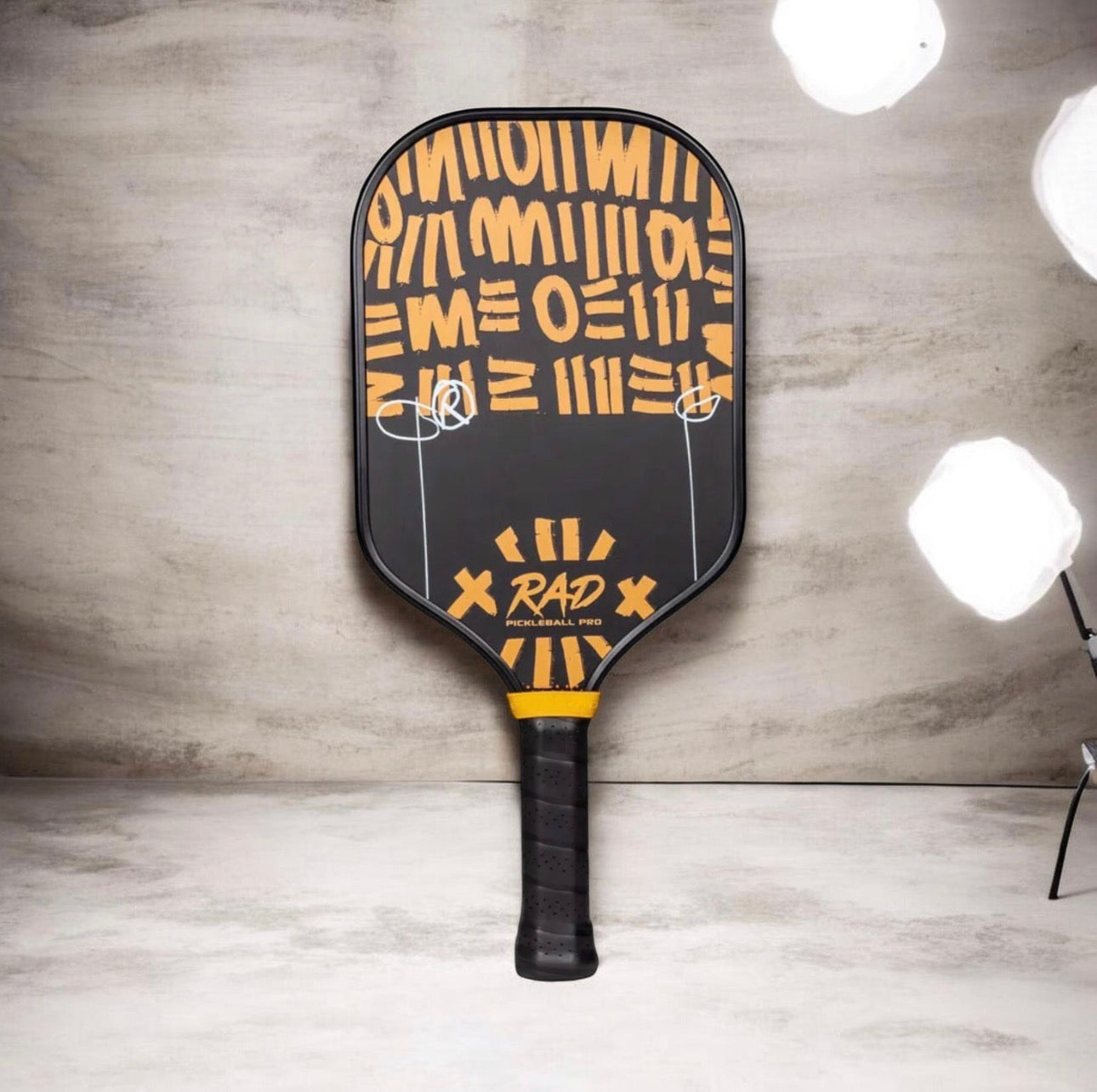 A black pickleball paddle with a yellow graphic pattern and the brand name 'RAP' printed on the face, standing upright against a gray textured wall with studio lights visible in the background.