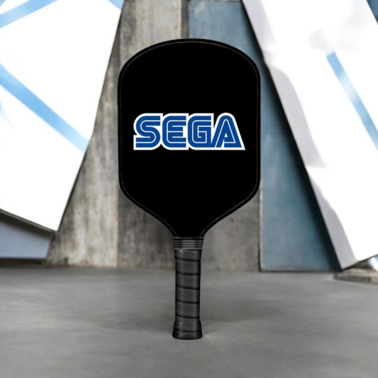 A black pickleball paddle with a textured black grip and the blue and white SEGA logo on the paddle face, standing upright on a gray concrete surface.