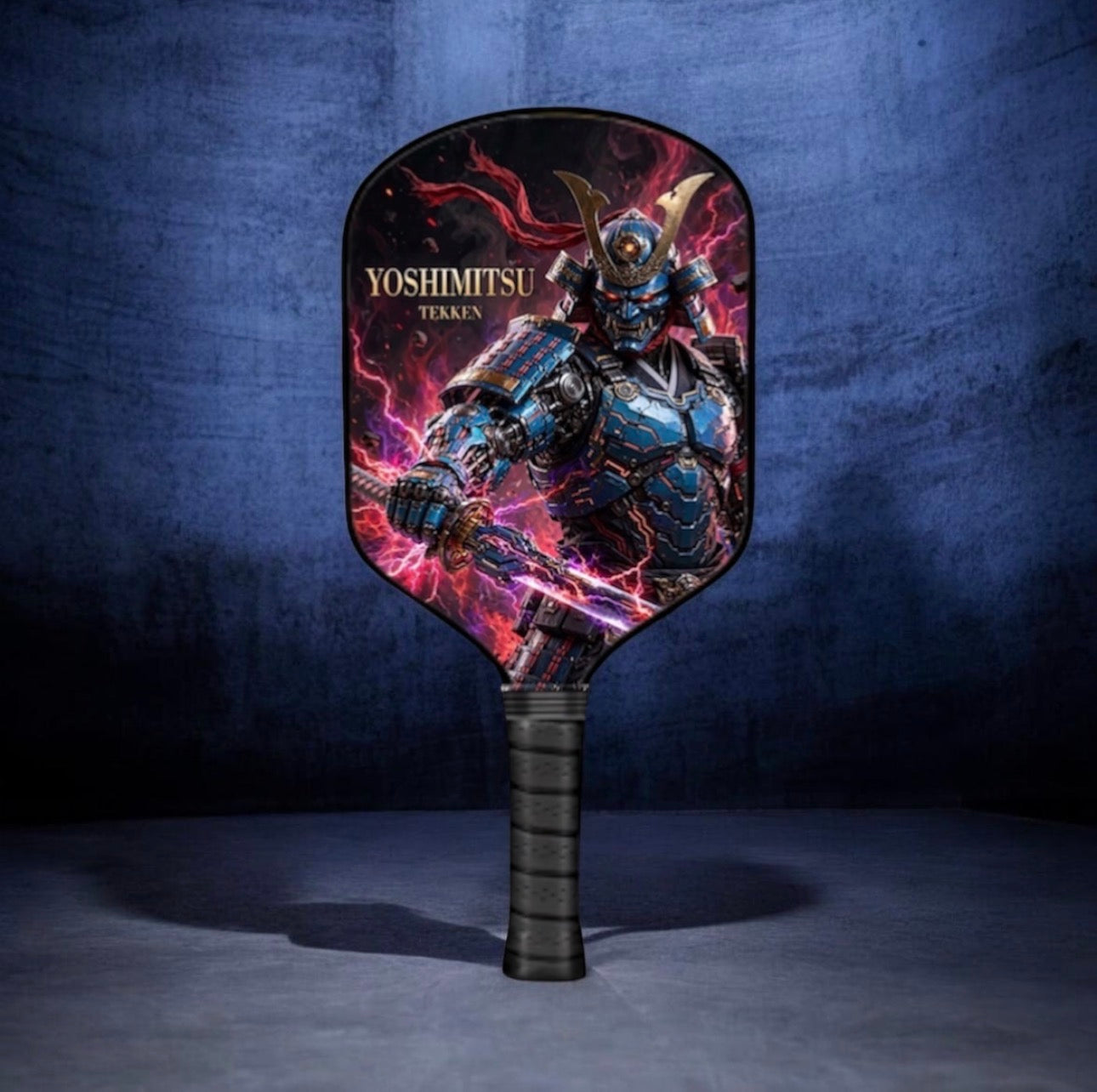 A pickleball paddle featuring a graphic print of a samurai warrior in blue and black armor with red and purple energy effects, and the text 'YOSHIMTSU TOKYO'. The paddle has a black wrapped handle and is set against a dark blue textured background.
