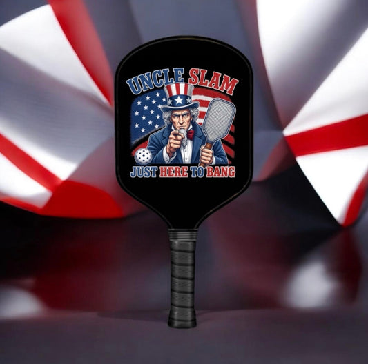 A black pickleball paddle featuring a patriotic Uncle Sam graphic with the text 'UNCLE SLAM JUST HERE TO BANG' in red, white, and blue, set against a blurred American flag background.