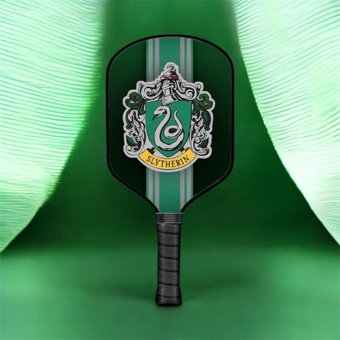 A pickleball paddle featuring the Slytherin crest from Harry Potter, with a dark green and light green striped design and a black wrapped handle, set against a green background with draped fabric.