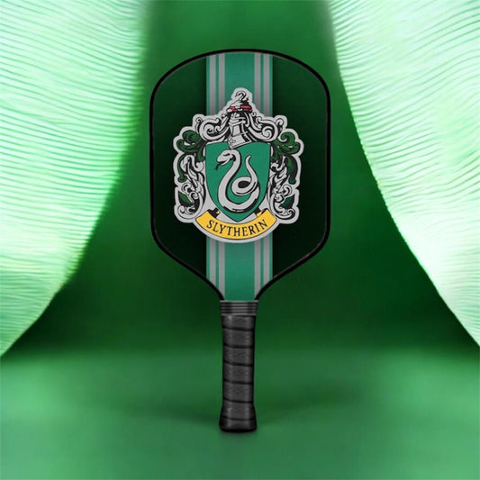 A pickleball paddle featuring the Slytherin crest from Harry Potter, with a dark green and light green striped design and a black wrapped handle, set against a green background with draped fabric.